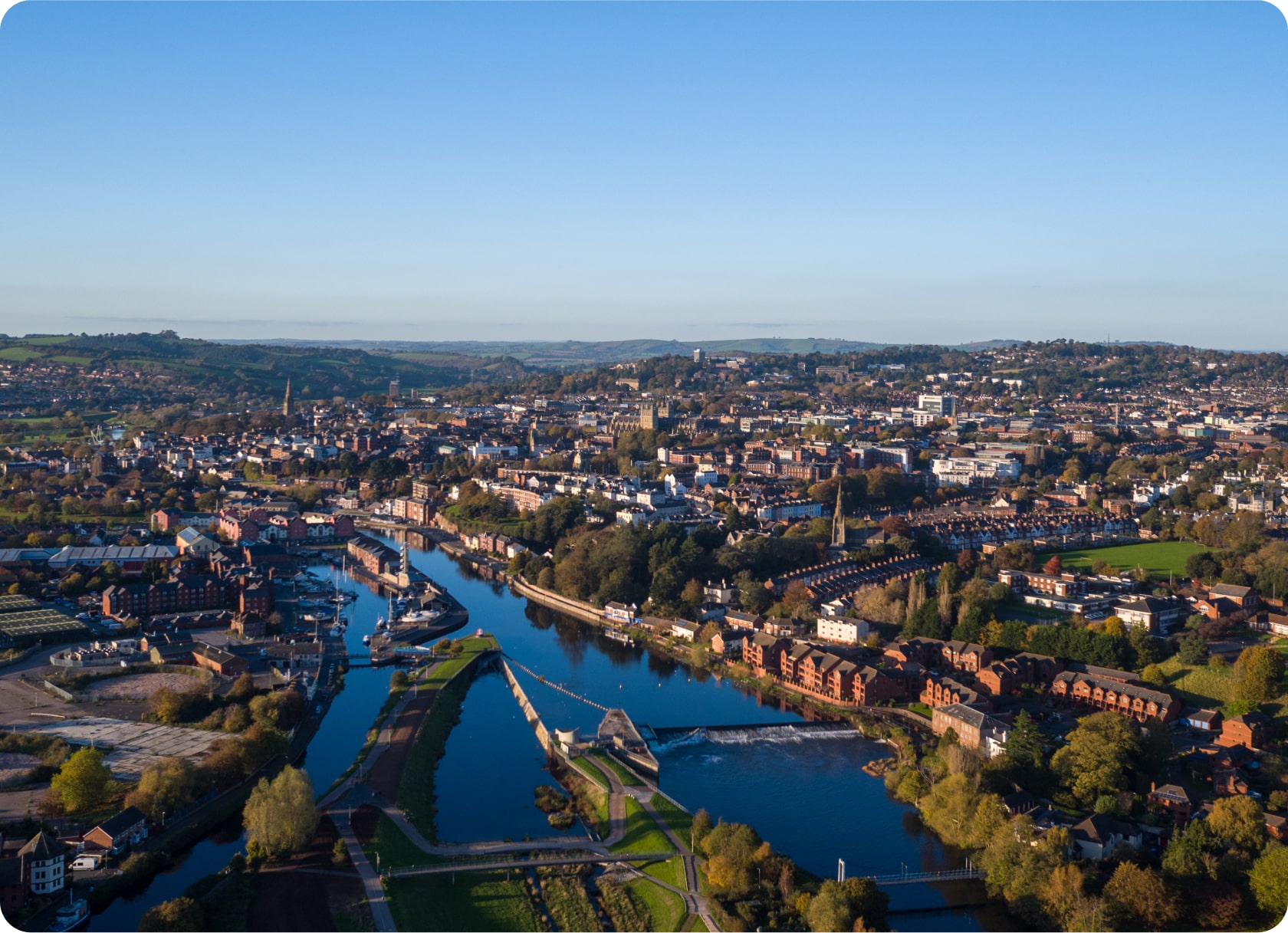 Aerial view of a city with a river running through its center. The landscape features a mix of urban and green areas, with numerous buildings, bridges over the river, and patches of trees. The horizon shows distant hills under a clear blue sky.