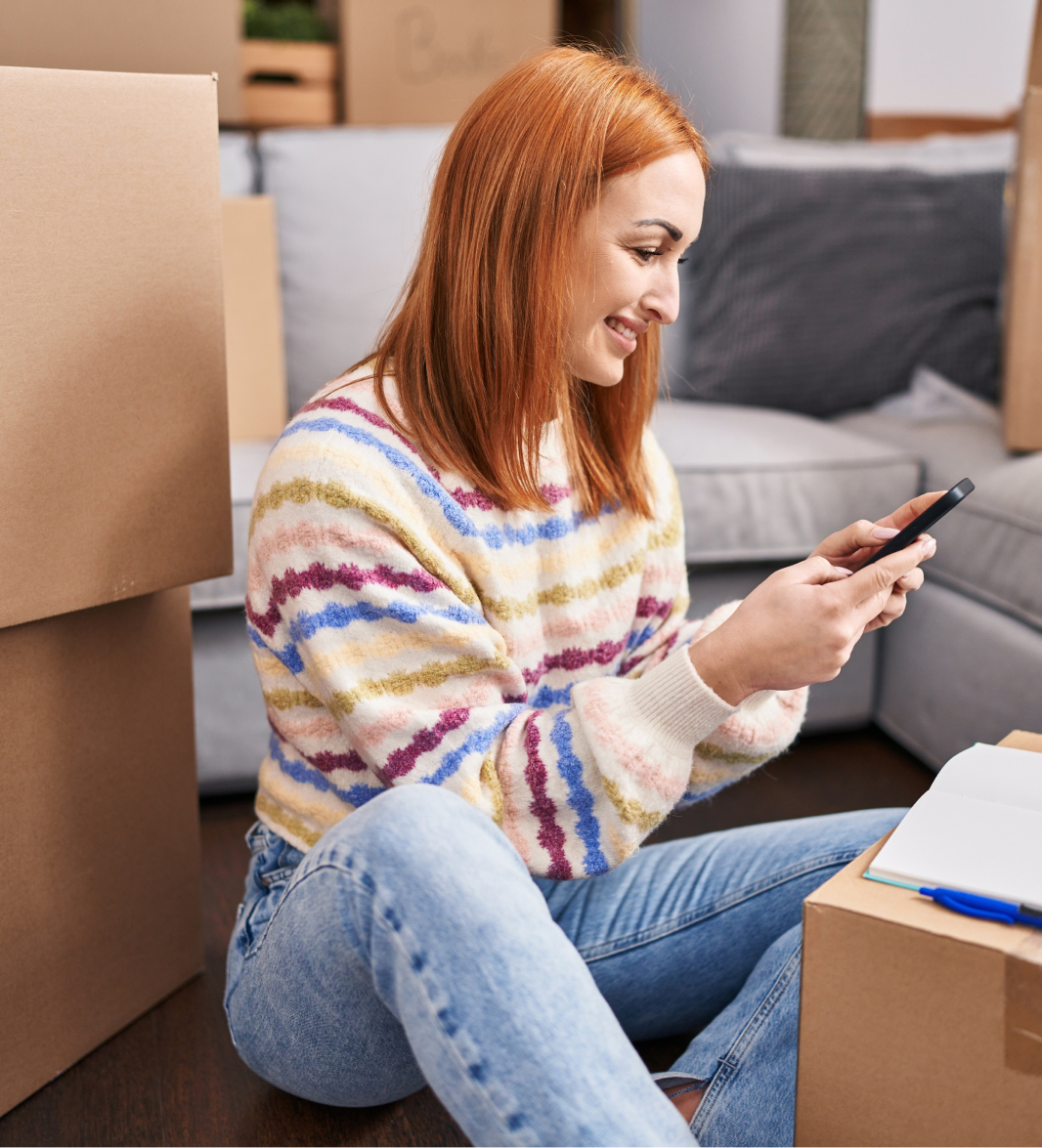 A woman with red hair, wearing a colorful striped sweater and blue jeans, sits on the floor among unpacked cardboard boxes. She is smiling and looking at her smartphone. An open notebook is placed on one of the boxes nearby.