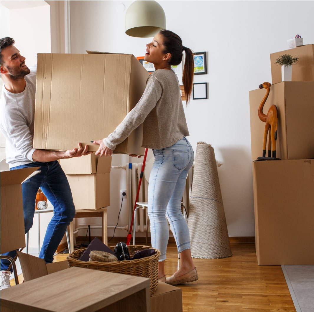 A man and a woman are carrying a large cardboard box together in a room filled with various packed boxes and household items. The room appears to be in the process of being packed or unpacked. The floor is wooden, and the atmosphere is busy.