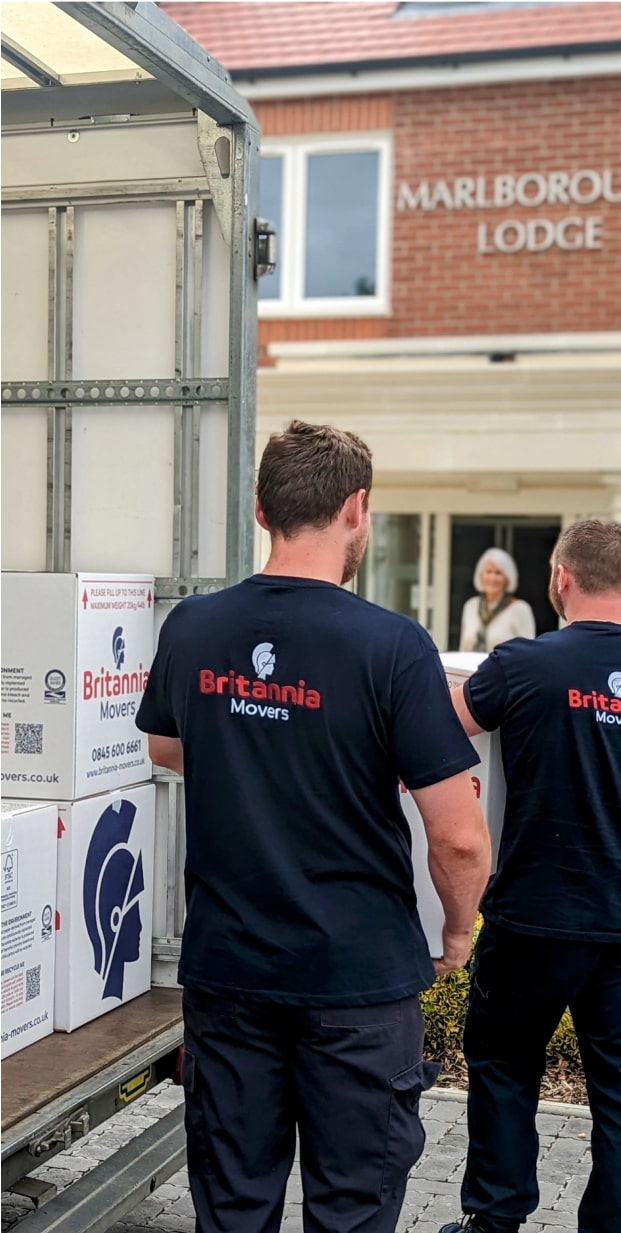 Two movers wearing Britannia Movers T-shirts are unloading boxes from a truck. The truck's back is visible, and the boxes are branded with Britannia Movers logos. In the background, a smiling older woman stands at the entrance of a building labeled "Marlborough Lodge.