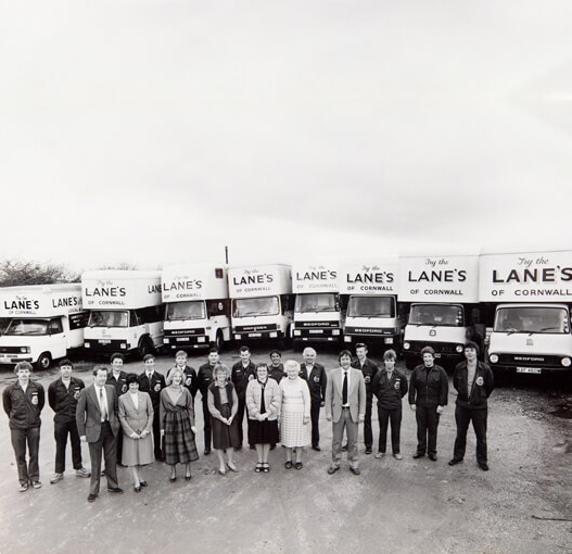 A group of 17 people stands in front of a line of delivery trucks with "Lane's of Cornwall" branding. The trucks are white with large logos on the sides. The people, dressed in business and casual attire, are arranged in a semi-circle facing the camera.