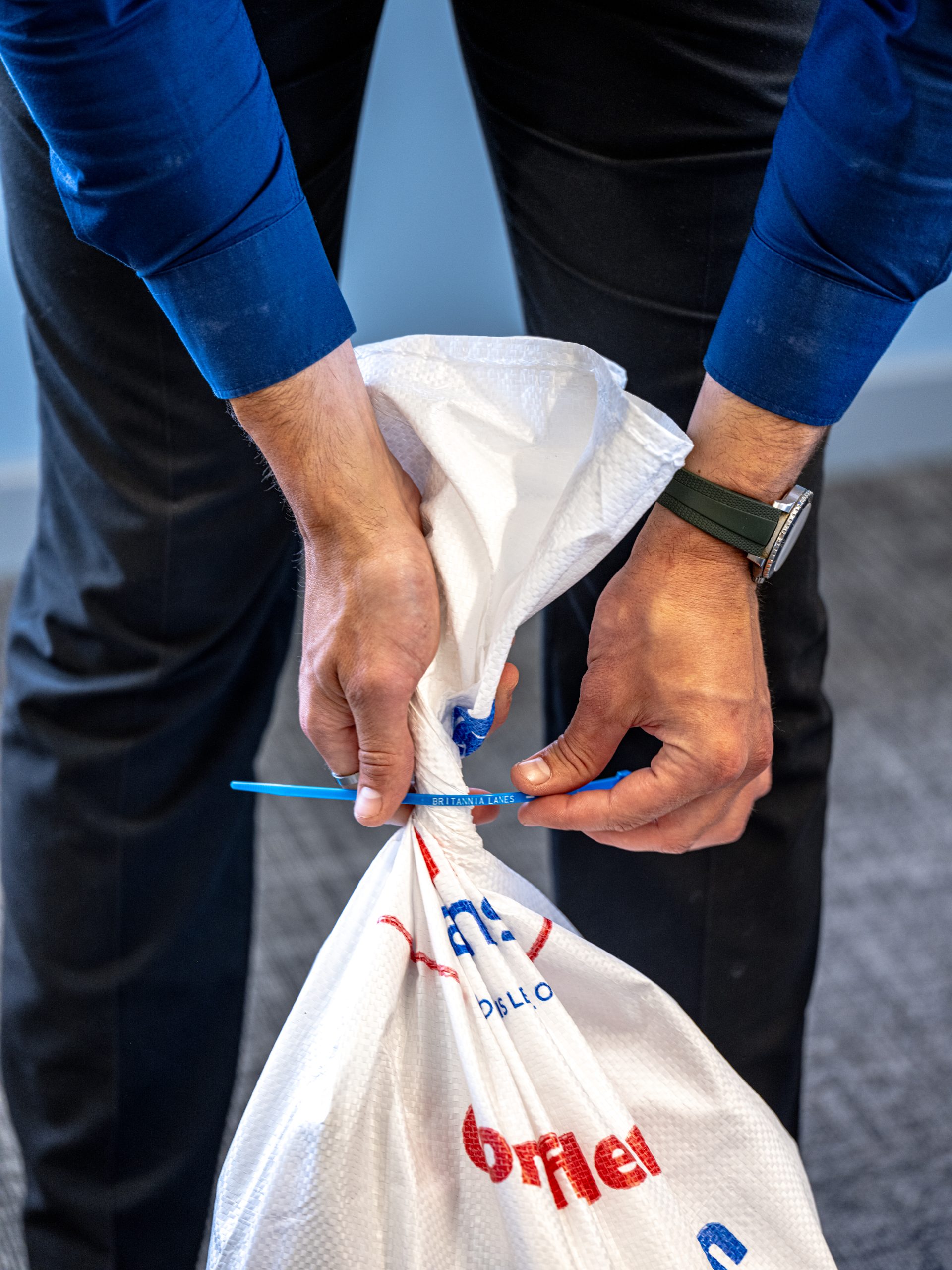 A person wearing a blue shirt and black pants ties a white sack with red and blue text on it using a blue zip tie. The person has a watch on their left wrist and is standing on a grey carpeted floor.