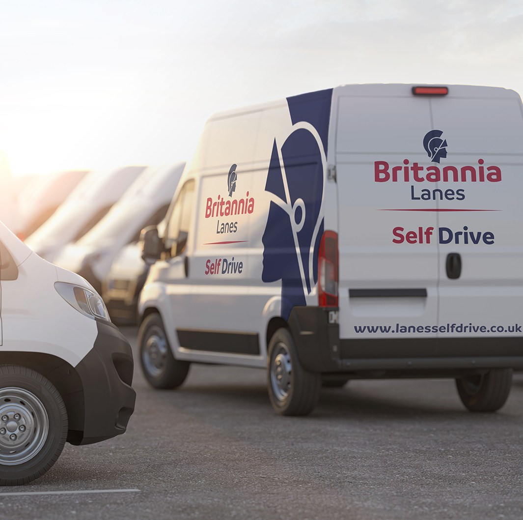 A row of parked white vans with "Britannia Lanes" and "Self Drive" branding. The nearest van has a prominent blue logo with a silhouette and the website www.lanesselfdrive.co.uk written on it. The sun is setting, casting a warm light across the scene.