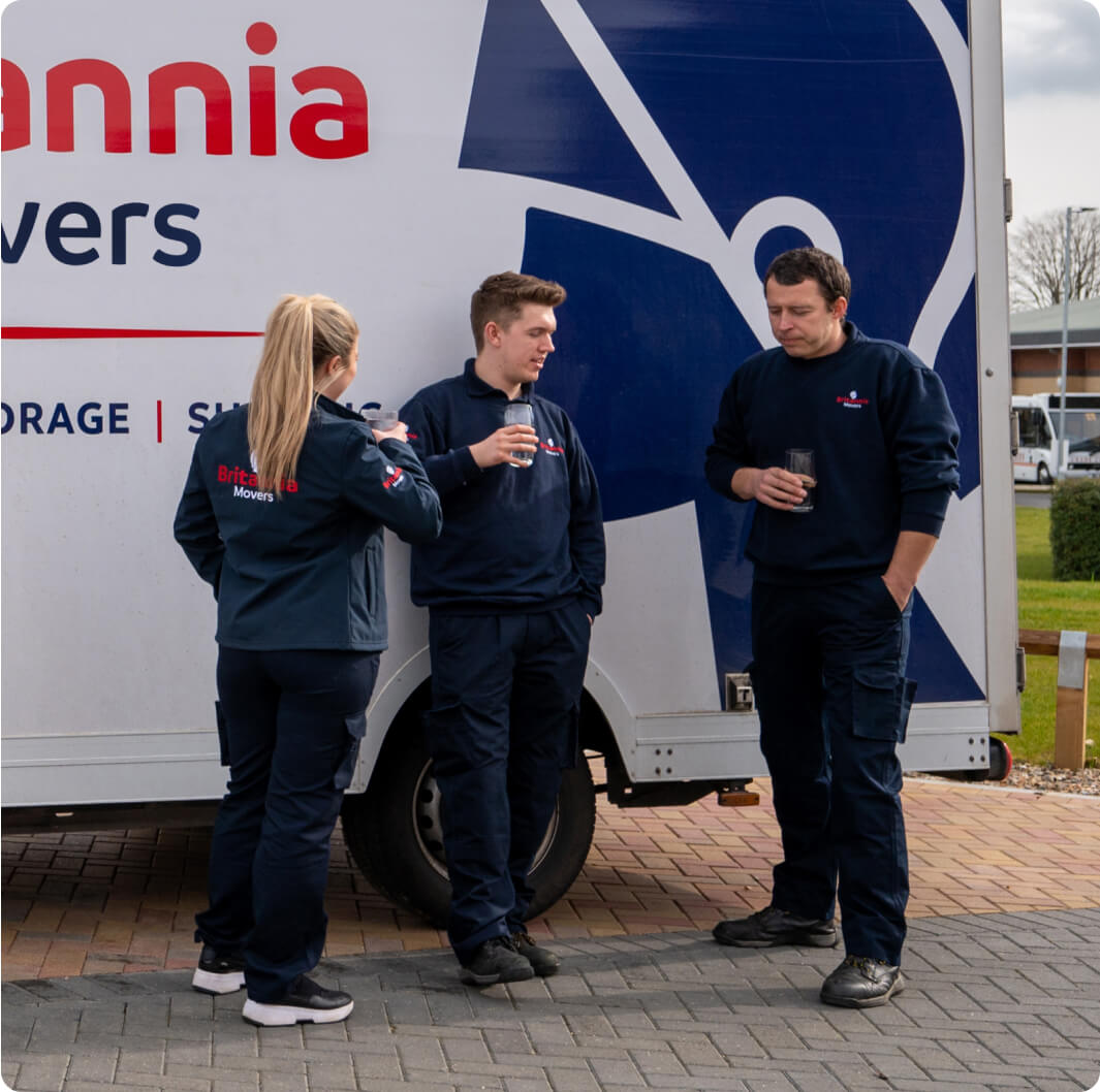 Three movers in uniform stand in front of a Britannia Movers truck, taking a break. Two of them hold drinks while conversing. The truck's sign displays "Britannia Movers" along with services like storage and shipping. They appear relaxed, enjoying their break.