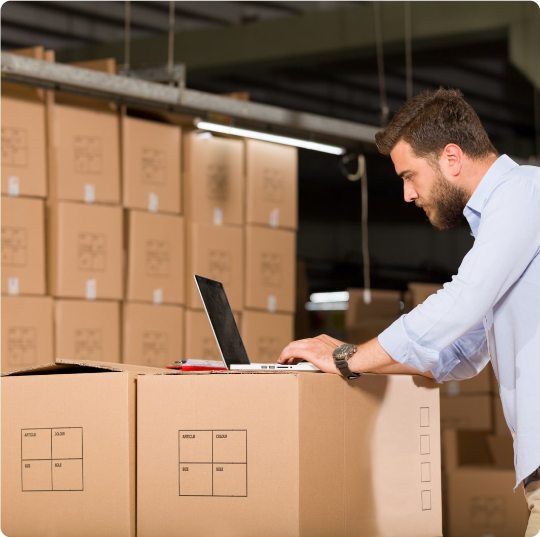 A man with a beard and wearing a light blue shirt is working on a laptop placed on top of cardboard boxes in a warehouse. The background is filled with stacks of similar boxes.