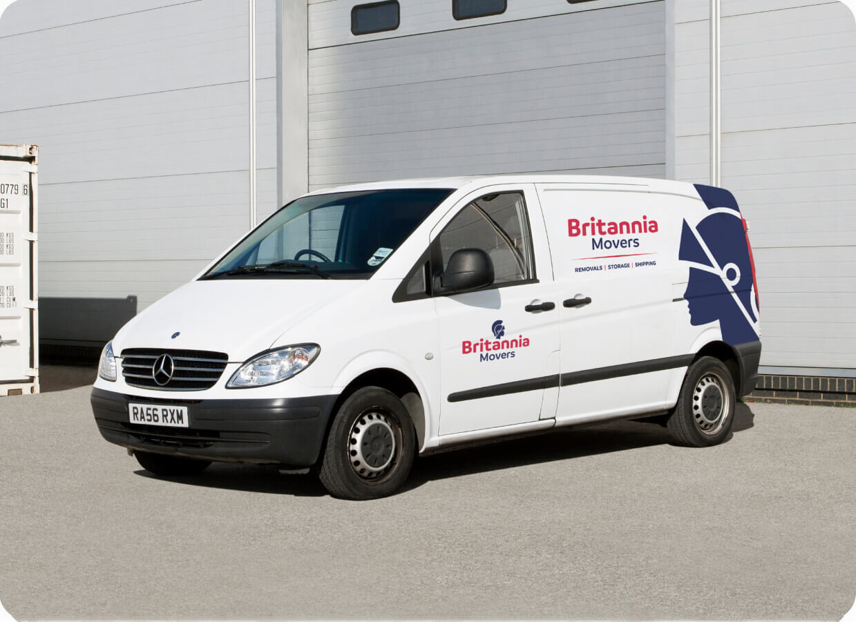 A white Mercedes van with "Britannia Movers" branding on the side and rear door is parked in front of a gray industrial building. The branding includes a blue and red logo with the text "Removals | Storage | Shipping" underneath. The van's license plate reads "RA56 RXM".