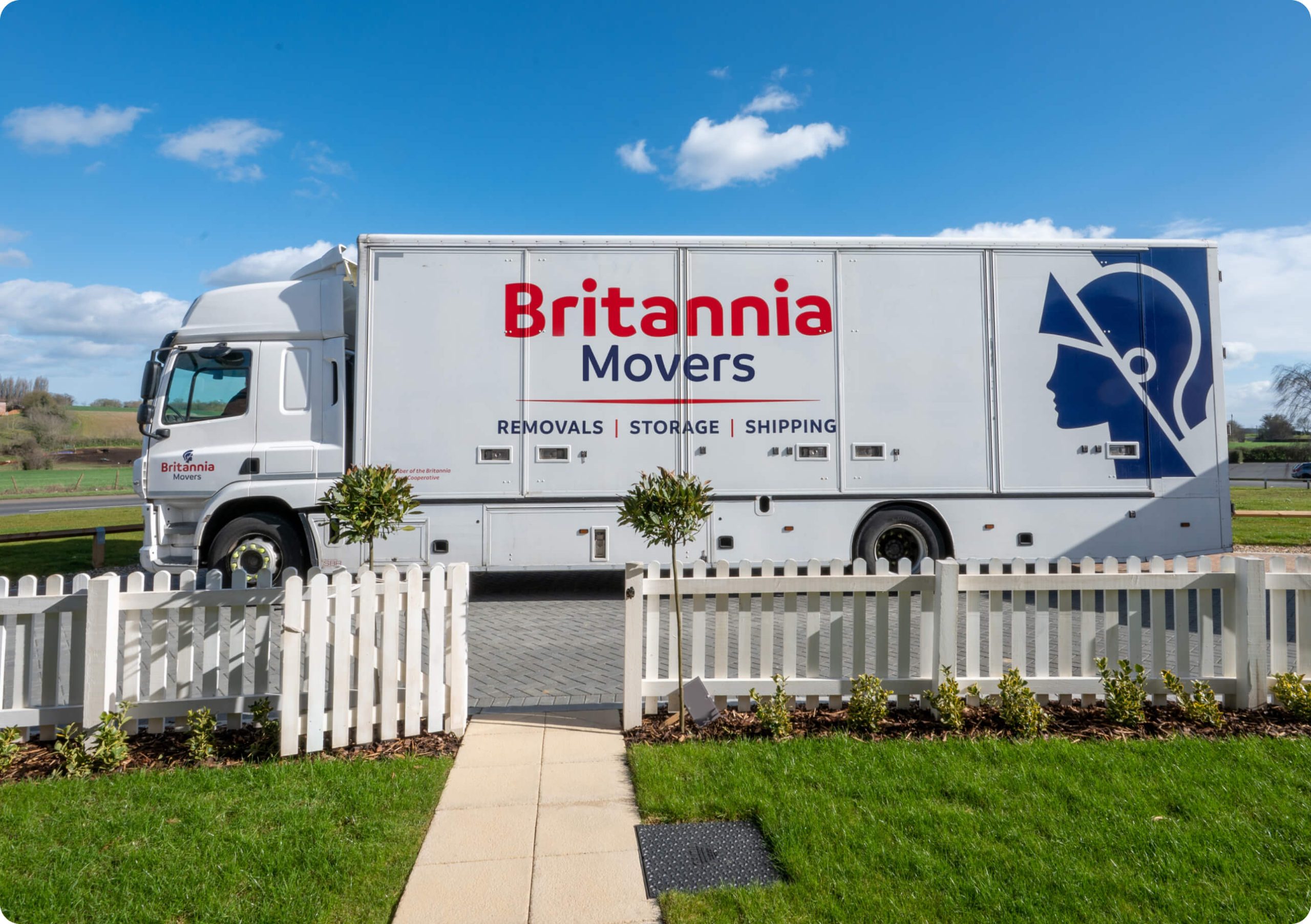 A large white truck with "Britannia Movers" and an image of a stylized face profile on the side is parked next to a white picket fence. The truck is labeled for removals, storage, and shipping services. The sky is clear and blue with a few clouds in the background.