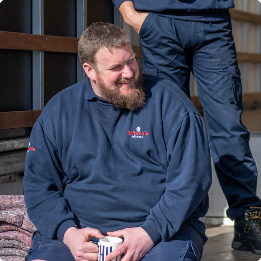 A bearded man in a blue Britannia Movers uniform sits with a striped mug, smiling. Another person in similar attire stands beside him, but their face is not visible. They appear to be outdoors near moving equipment.