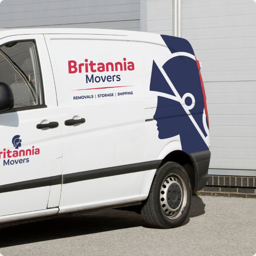 A white van with the "Britannia Movers" logo in red and blue on its side. The logo includes a profile of a blue helmeted figure on the rear side of the van. The text below the company's name reads: "REMOVALS | STORAGE | SHIPPING." The van is parked in front of a light-colored building.