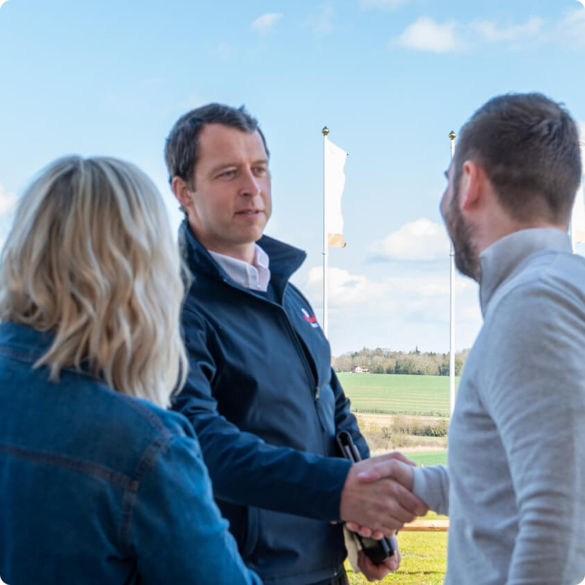 A man in a navy jacket shakes hands with another man wearing a grey shirt. A woman with blonde hair and a denim jacket stands beside them. They are outdoors with a clear blue sky and fields in the background. The image captures a friendly interaction.