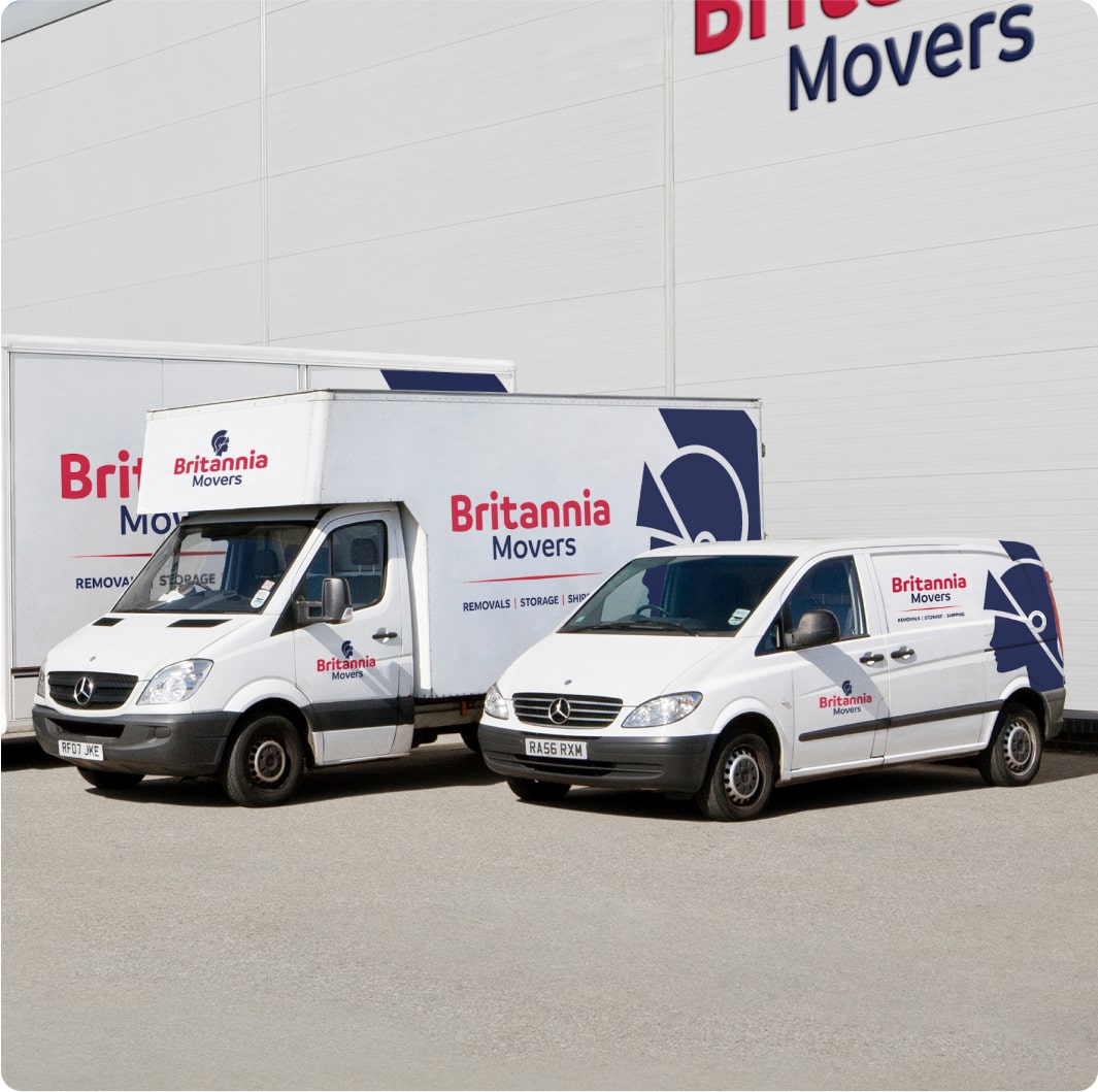 Two white delivery vans, branded with "Britannia Movers" logo and text, are parked next to a large white building with "Britannia Movers" signage. The larger van on the left and smaller van on the right both feature the company name and graphic.