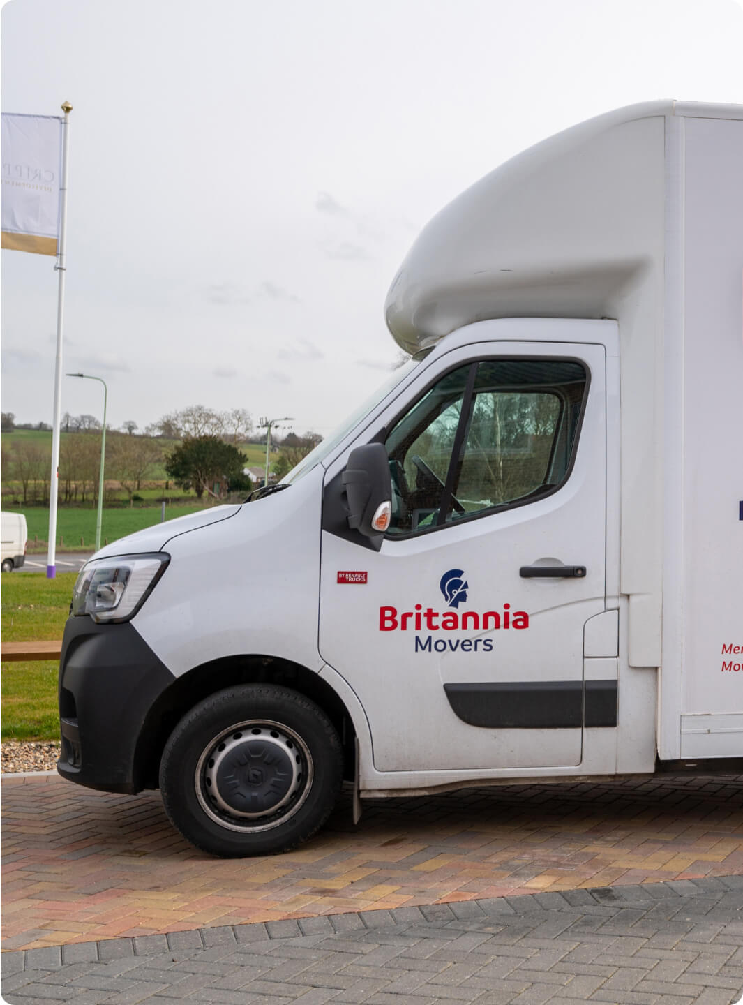 A white moving van with the logo "Britannia Movers" on the door is parked on a brick driveway. The van is positioned facing to the right, and the background includes a grassy field, a flagpole with a white flag, and some trees.