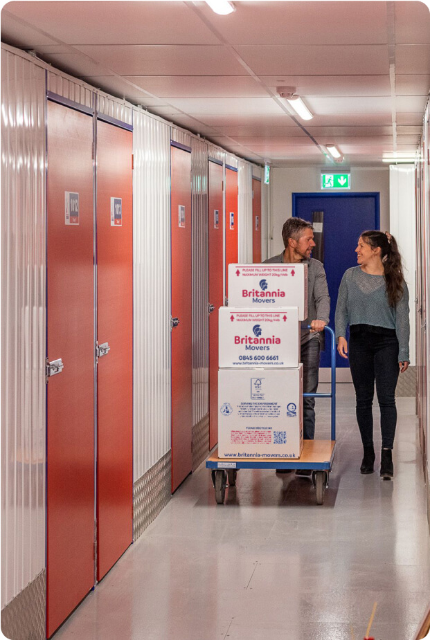 A man and a woman are moving cardboard boxes labeled "Britannia Movers" on a blue hand truck through a hallway lined with red storage unit doors. They are both smiling and appear to be having a conversation. The woman is holding the handle of the cart and the man is pulling it from the front.